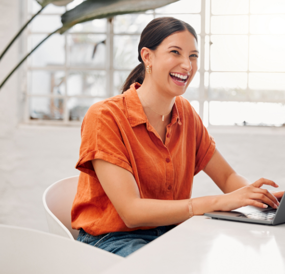 A woman in an orange top smiling while typing on her laptop, representing JTF Marketing’s expertise in creating effective landing page templates.